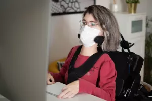 Disabled employee with muscular dystrophy at the office working in front of a computer wearing a surgical mask.