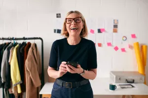 Laughing mature business owner with mobile phone in front of desk in fashion start-up.