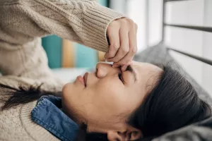 Patient lying on bed in home and pinching top of nose between eyes to relieve migraine.