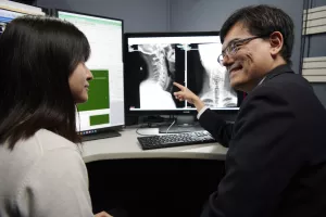 Radiologist teaching a resident by showing and talking about a scan of a patient's head, neck, spine and shoulders on the computer screen.